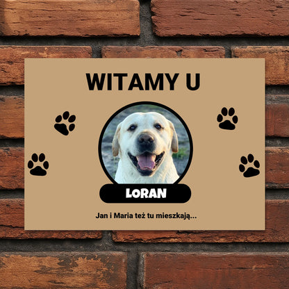 Tabliczka powitalna z napisem „Witamy u” i zdjęciem psa w okrągłej ramce.