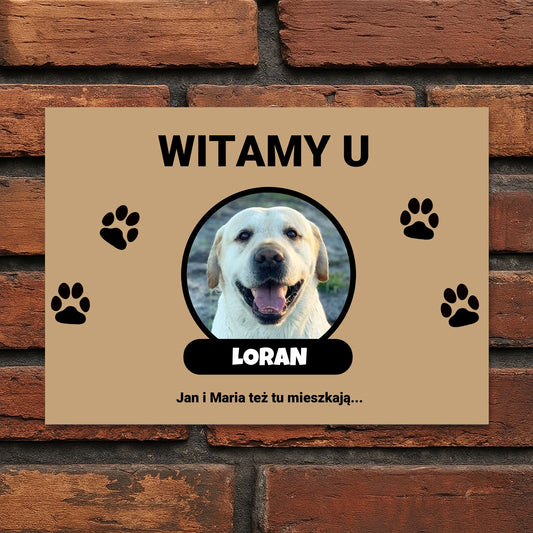 Tabliczka powitalna z napisem „Witamy u” i zdjęciem psa w okrągłej ramce.