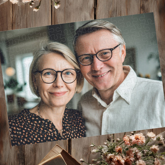 Puzzle z jednym dużym zdjęciem uśmiechniętej pary seniorów w okularach, z widocznymi liniami puzzli, ułożone na drewnianym stole z kwiatami